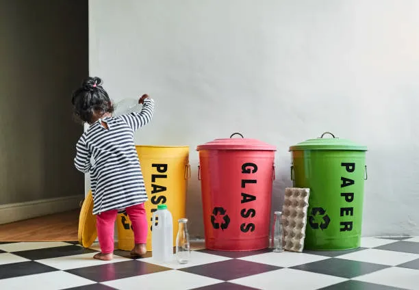 child in front of three plastic garbage bin that read plastic glass and paper 